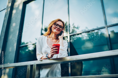 Smiling hipster girl in eyewear making phone call during break holding coffee takeaway standing on urban settings background, young woman talking on mobile with friend discussing plans for leisure