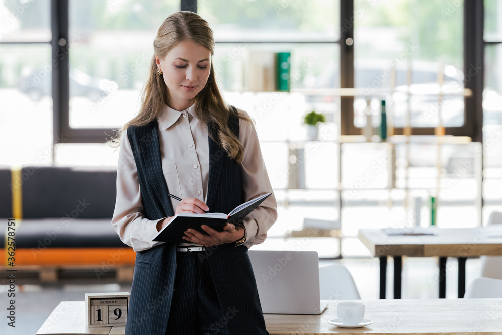 attractive businesswoman standing near table writing in notebook
