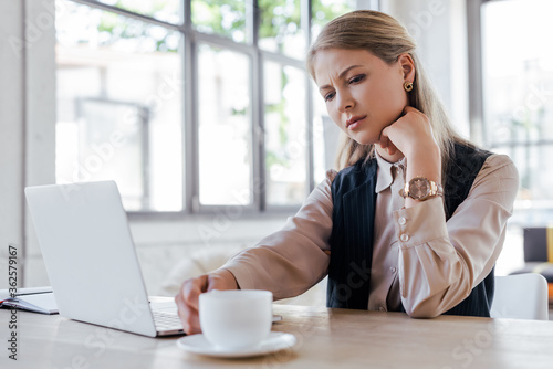selective focus of exhausted businesswoman touching cup of coffee near laptop