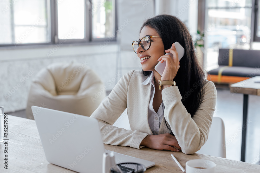 selective focus of happy businesswoman in glasses talking on smartphone near laptop selective focus of happy businesswoman in glasses talking on smartphone near laptop