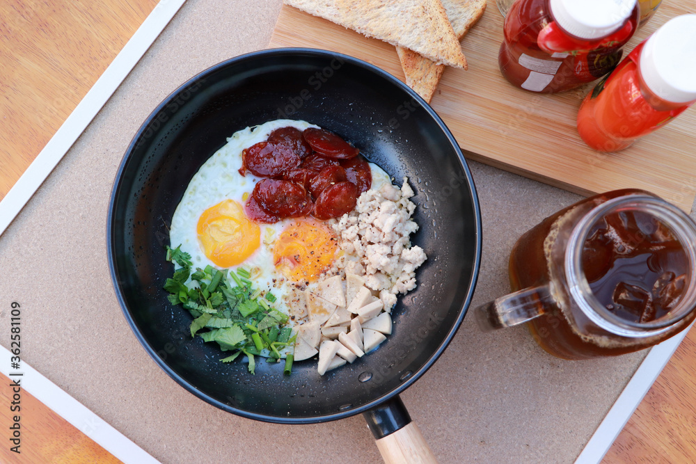 pan fried egg with toppings, Chinese sausage and pork sausage in the black pan with flavoring, slice of bread and water. pan fried egg with toppings, Chinese sausage and pork sausage in the black pan with flavoring, slice of bread and water.