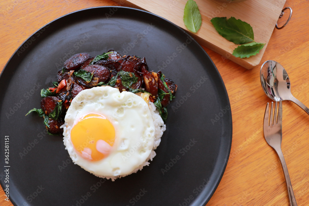 Chinese sausage fried with basil and fried egg with rice in the black round dish, Cooking ingredients with spoon and fork. Chinese sausage fried with basil and fried egg with rice in the black round dish, Cooking ingredients with spoon and fork.
