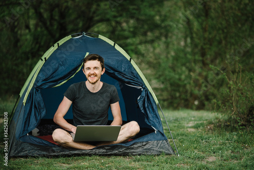 Man working on laptop in tent in nature. Young freelancer sitting in camp. Relaxing in camping site in forest, meadow. Remote work, outdoor activity in summer. Happy male relaxing, work on vacation.