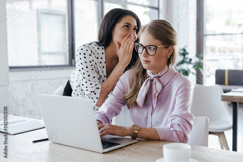 businesswoman whispering in ear of coworker in glasses while gossiping in office