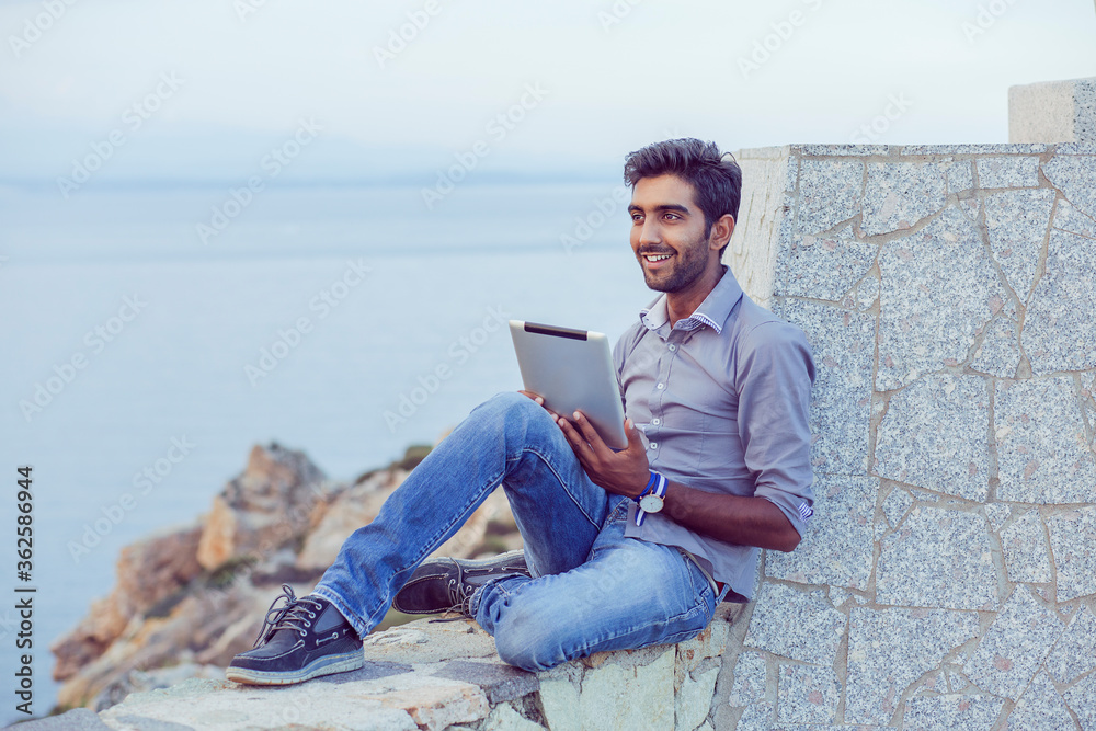 Man smiling happy while using pad sitting on a bridge above the sea