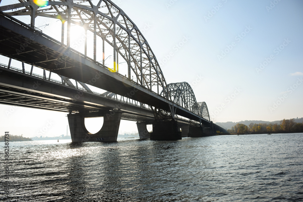 A long arched iron bridge over a wide river. A sun breaks through it.
