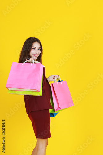 Beautiful woman in red suit smile and hand holding colorful shopping paper bags isolated on yellow background.