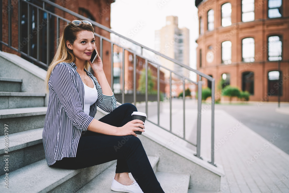 Fototapeta premium Young caucasian woman with nice smile talking on phone in pleasant atmosphere outdoors sitting on university stairs and having takeaway coffee, contemporary technology and communication concept