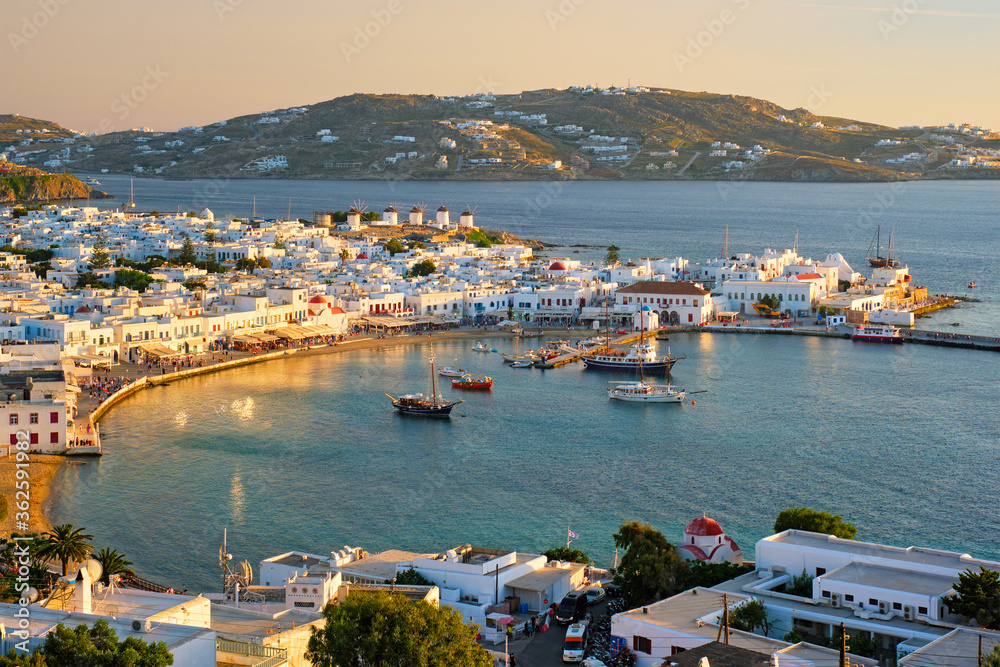 View of Mykonos town Greek tourist holiday vacation destination with famous windmills, and port with boats and yachtson sunset . Mykonos, Cyclades islands, Greece. With horizontal camera panning
