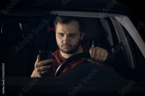 Unshaven confident serious young millenial male driver sits at steering wheel of car at night tired looking at smartphone worried concerned about problems. safe driving concept. Loneliness Concept.