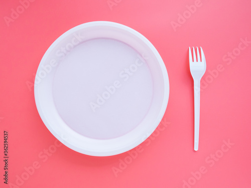 White plastic plate and fork on pink background, flat lay, top view. Bright minimalistic concept of eating and table setting. Bright slide to your presentation. The problem of disposal of plastic