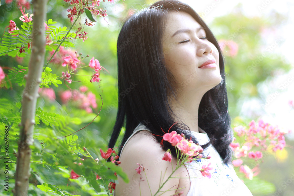Naklejka premium Beautiful asian woman in white dress close her eyes and smelling in blooming flowers with natural in garden sunlight. Thai or Chinese Girl enjoy on holiday