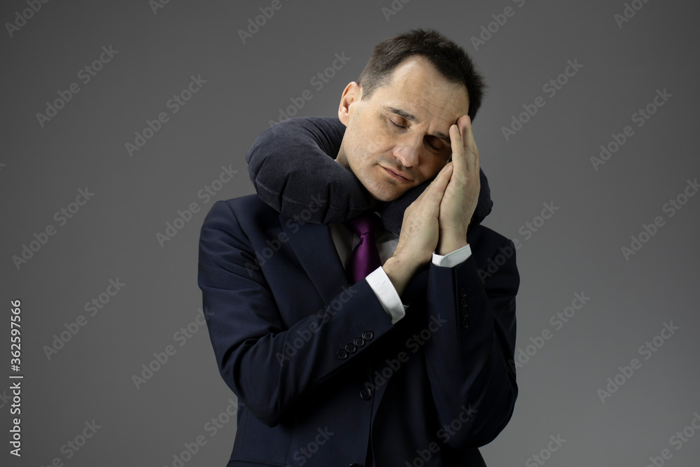 tired Exhausted middle-aged businessman in suit asleep standing up with his hands under his head with inflatable pillow around his neck isolated on gray. Fatigue and overload concept. tired Exhausted middle-aged businessman in suit asleep standing up with his hands under his head with inflatable pillow around his neck isolated on gray. Fatigue and overload concept.