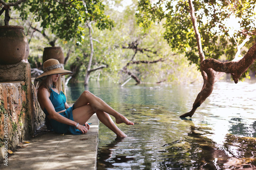 beautiful blonde woman in summer hat and aquamarine swimsuit posing near nature laguna in Mexico in summertime at green forest background 