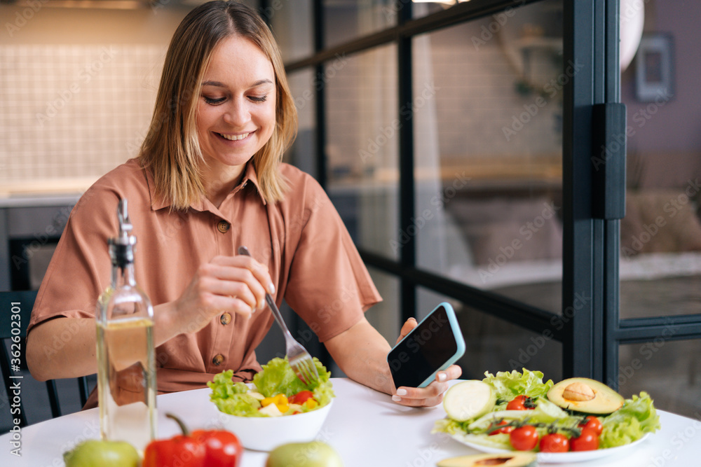 Young smiling woman mixing fresh vegetable salad while sitting at table in kitchen with modern interior, mobile phone on hand. Concept of healthy eating.
