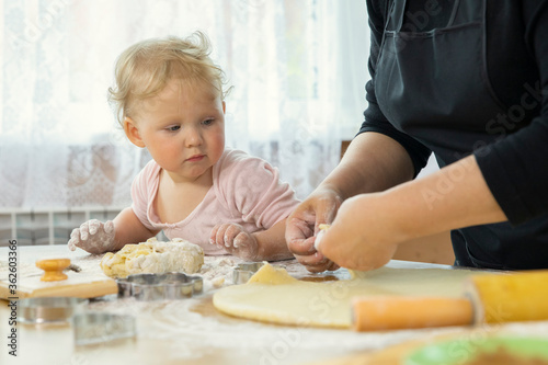 Close up view of little cute baby and grandma in apron cooking on kitchen, make homemade pastry. Grandmother and granddaughter preparing cookies for christmas. Child in flour teaching to bake