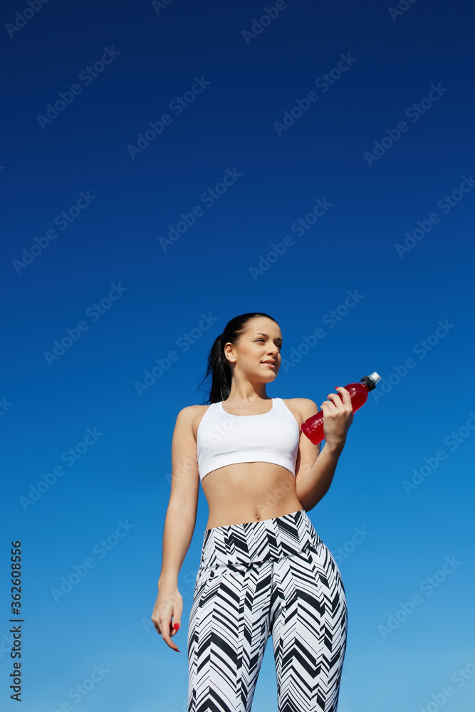 Healthy young woman taking a break after workout outdoors, athletic female standing on blue sky background while holding bottle of energy drink, fitness training at sunny afternoon in summer Healthy young woman taking a break after workout outdoors, athletic female standing on blue sky background while holding bottle of energy drink, fitness training at sunny afternoon in summer