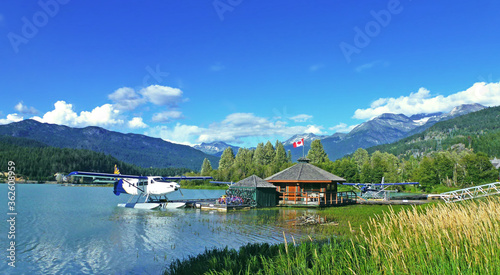 Whistler - Green Lake Landscape. The view on the boat house with Canadian national flag and harbor plane parked on the lake. Pine trees and mountains in the background.