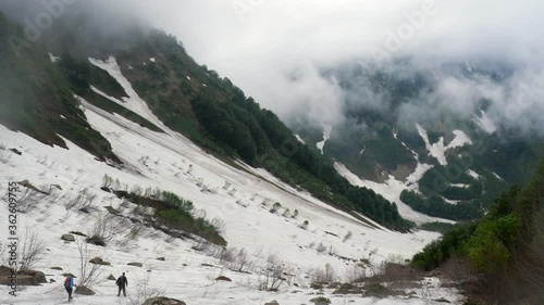 Snow-covered mountainside. Valley under the snow. Mists walk in the distance. Forest on the slopes around. Silhouettes of two tourists walking along a high-mountainous snowfield.