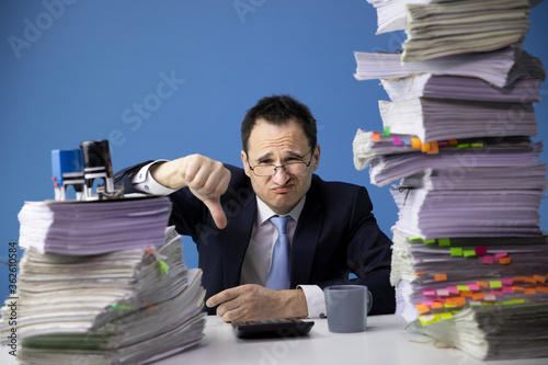 businessman in glass and tie shows finger down sitting at office desk with huge stack of documents looking sad and depressed in business stress and crisis concept. Fatigue and overload concept.