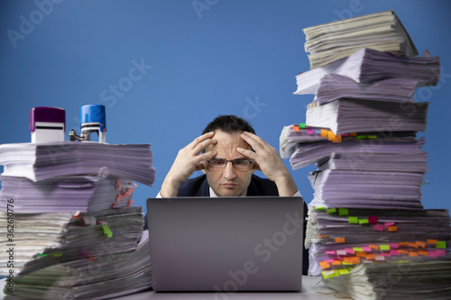 Tired concerned businessman in glass and tie sitting at office desk with huge pile of documents working on computer laptop looking sad and depressed in Fatigue and overload concept. isolated on blue