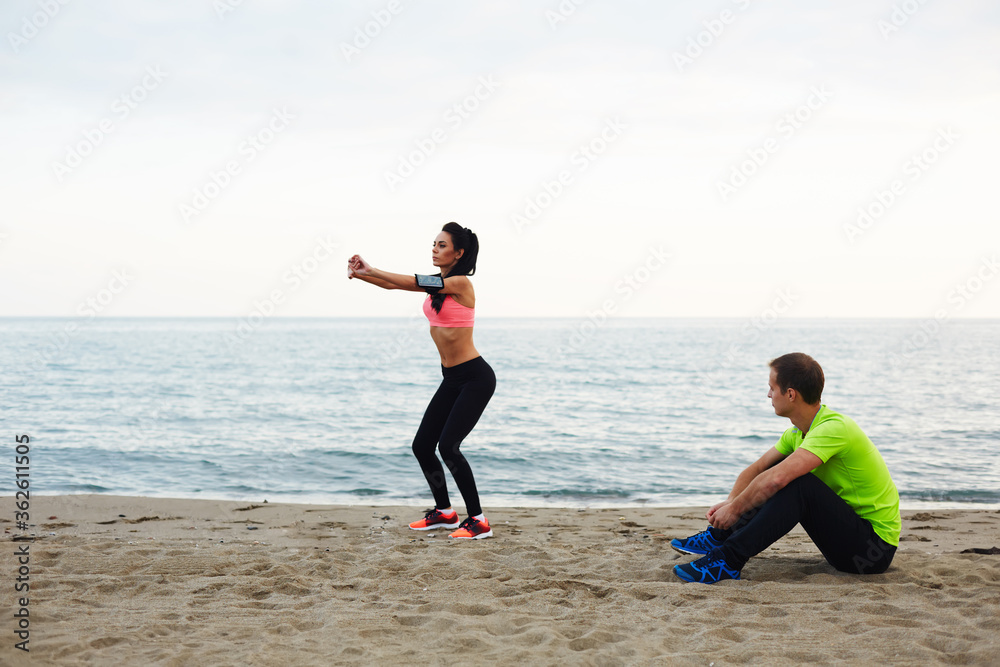 Personal trainer observing how young woman doing squat exercises, fitness couple training on the beach against sea at evening time.