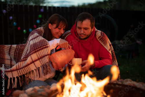 happy couple in love at picnic with bonfire. Handsome Man And Beautiful Woman pouring coffee from thermos into cup at romantic party by campfire. togetherness concept