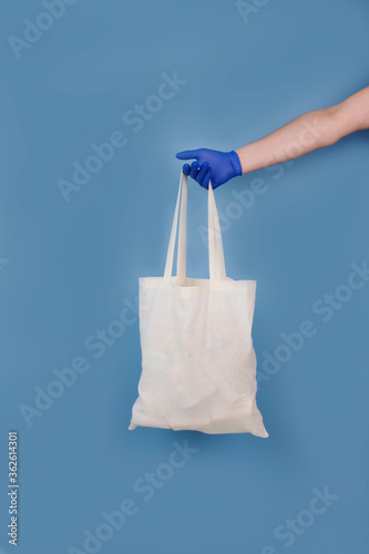Vertical composition with man's hand in blue disposable protective gloves, holding white bag with food or goods donations on blue trendy background. Delivery or donation concept, mockup and copyspace