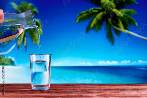 A glass full of drinking water on a wooden table over natural blue sea beach and palm trees