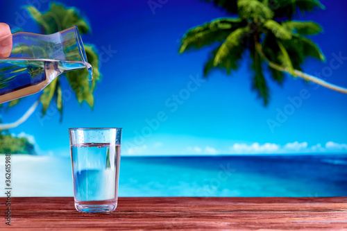 A glass full of drinking water on a wooden table over natural blue sea beach and palm trees
