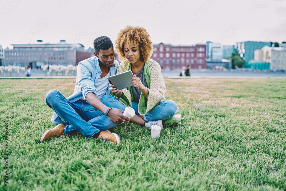 Happy couple in love spending time together in park on green grass and watching video online on modern touch pad device.Positive african american young man and woman doing shopping together on tablet Happy couple in love spending time together in park on green grass and watching video online on modern touch pad device.Positive african american young man and woman doing shopping together on tablet