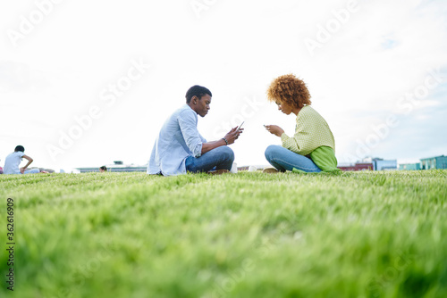 Addicted african american friends bloggers ignoring live communication while chatting online in social networks on smartphone using 4G internet sitting against each other on green grass in park