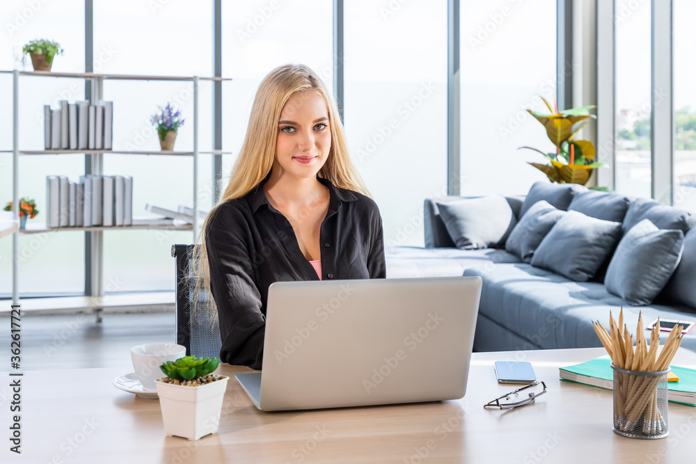 Portrait of smiling young Caucasian woman with blonde hair freelancer working at home, sitting at desk in living room using typing laptop computer; looking at camera Portrait of smiling young Caucasian woman with blonde hair freelancer working at home, sitting at desk in living room using typing laptop computer; looking at camera