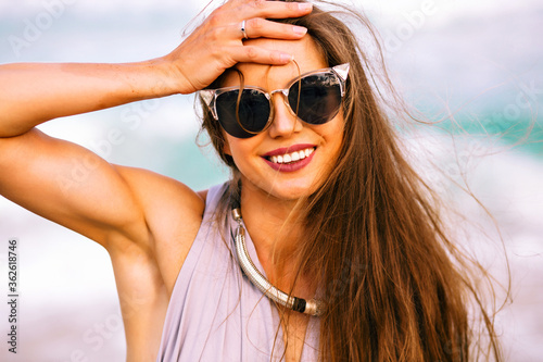 Close up cheerful portrait of pretty elegant woman posing near ocean , wearing trendy glamour sunglasses and necklace, long hairs and big smile, natural summer beauty.