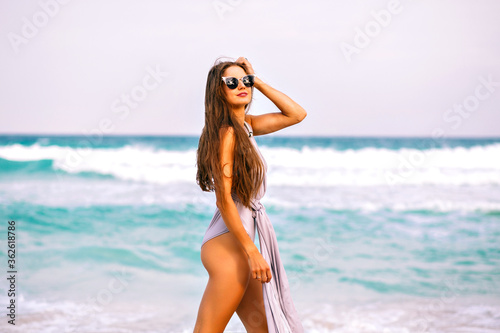 Summer portrait of sensual pretty woman posing near ocean, long hairs, luxury trendy bikini and accessories, vacation time. Walking alone, perfect sportive tanned body, blue ocean.