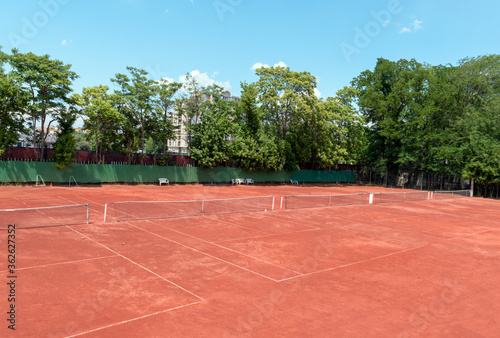 Empty tennis court on sunny summer day. View from above of a red clay tennis court, green trees and a blue sky in the background. Outdoor sports playground for tennis. Copy space for text or design