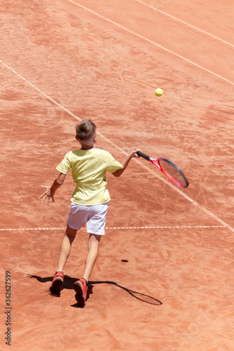 Child boy plays tennis on clay court. Young male tennis player hits the ball in flight in action. Kids tennis concept. Sports action frame. Motion. Vertical banner with copy space © Elena