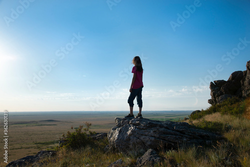 Girl with long hair stands on the edge of a high cliff above the green fields.