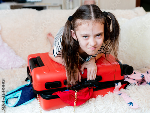 A little girl has Packed too many things on vacation, and can't close the red suitcase.