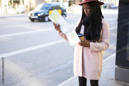 Pensive trendy dressed female traveler using map for searching right direction for destination standing on city avenue, young afro american international hipster girls searching location outdoors