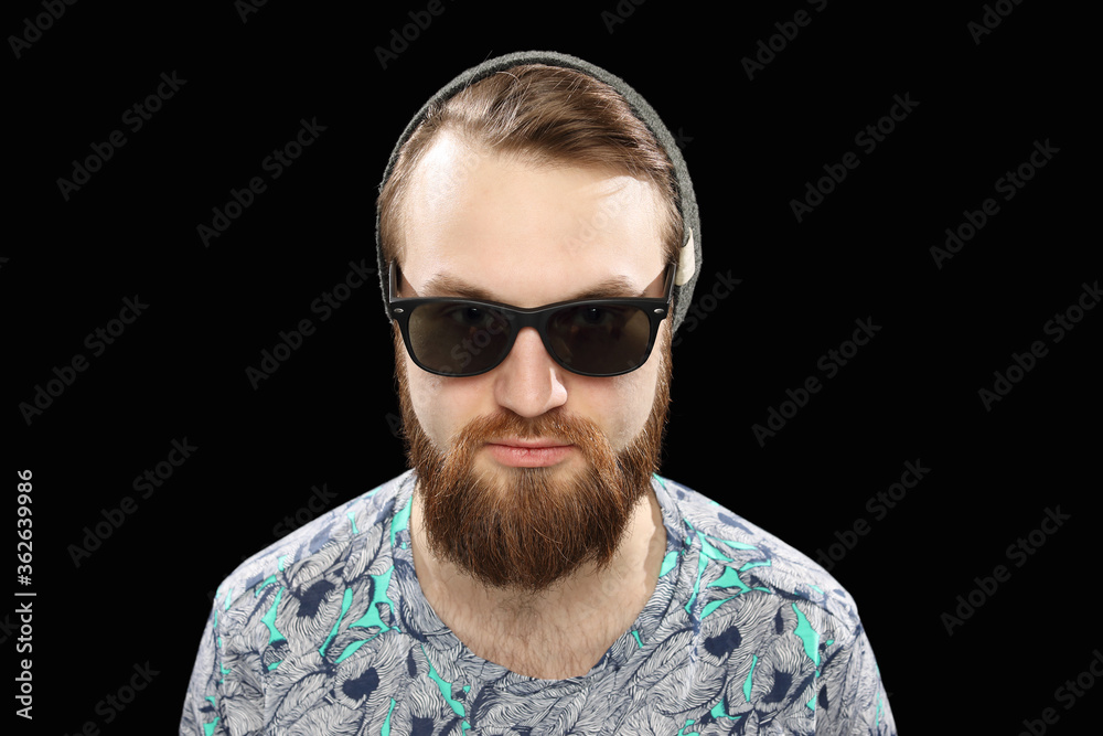 Portrait of young hipster man in sunglasses and hat posing on black background smiling bearded