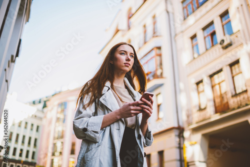 Gorgeous teenager dressed in trendy clothing looking away during walking at street. young hipster girl using application on smartphone connected to 4G and browsing interesting places to visit