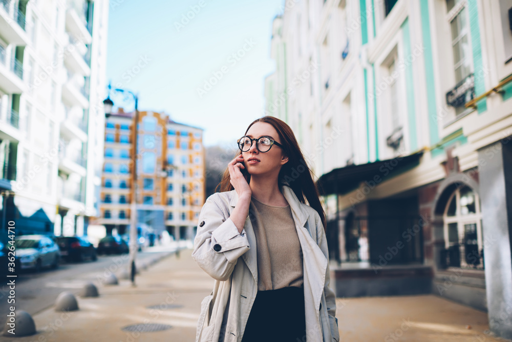 Thoughtful attractive female tourist walking during city tour enjoying spring weather and urban architecture communicating in roaming, hipster girl in trendy outfit arranging meeting talking on phone