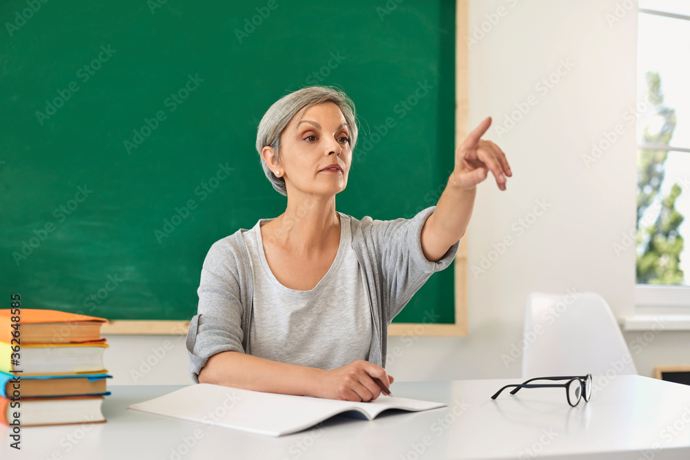 School education. Senior female teacher sitting at table and pointing somewhere in class. Mature educator during lesson School education. Senior female teacher sitting at table and pointing somewhere in class. Mature educator during lesson