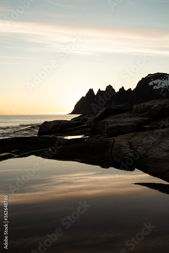 Amazing view on Senja island, Norway. Sunset light during polar day. Place called devil teeth - characteristic sharp mountains. Calm arctic ocean. No waves. Rocky beach. Reflection in water puddle.