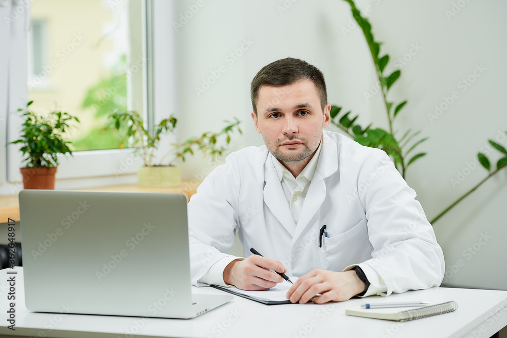 A caucasian doctor in a white lab coat is doing paperwork near a laptop in a hospital. A therapist is waiting for a patient in a doctor's office.