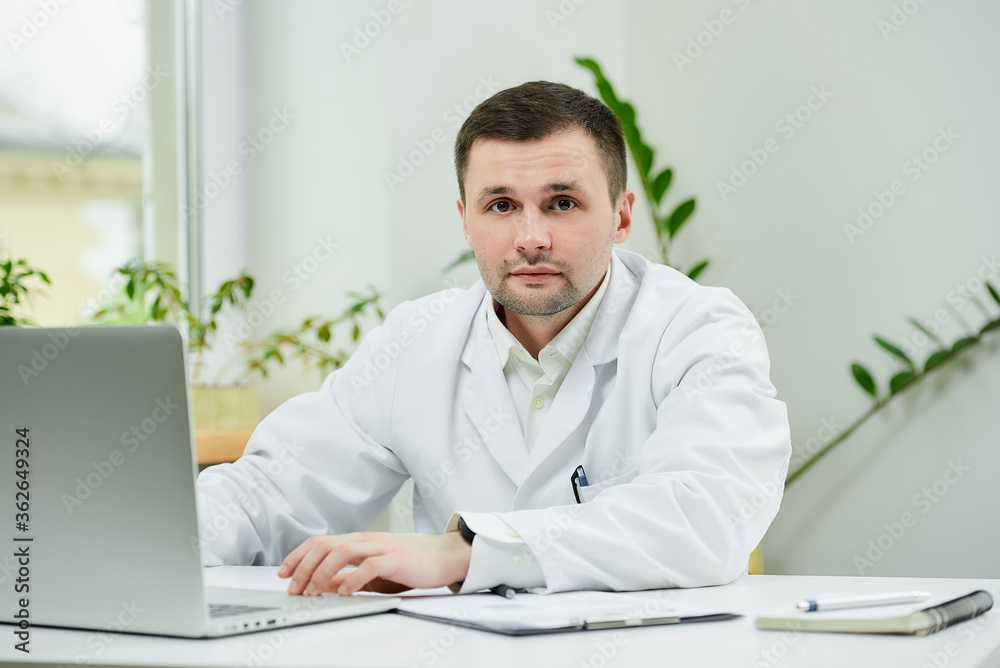 A serious caucasian doctor in a white lab coat was distracted while searching medical information on a laptop in a hospital. A therapist near a computer in a doctor's office.