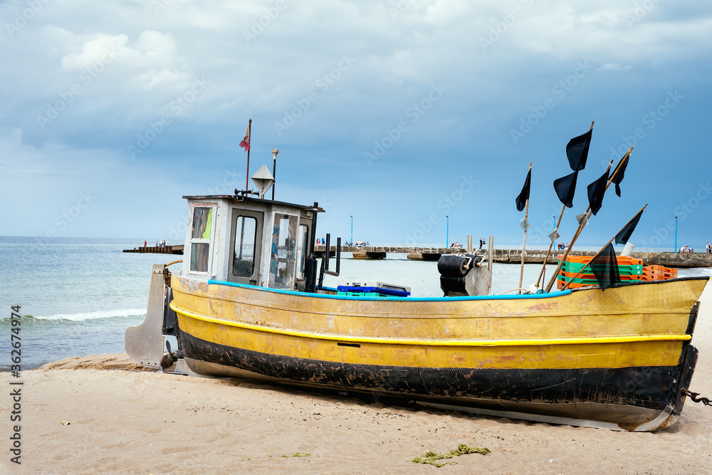 Fototapeta premium Old and rusty fishing boat and equipment on sandy beach, getting ready for fishing trip. Pier and blue sea in background, Niechorze, Poland