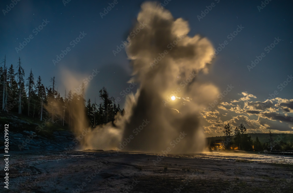 Moonlight eruption of a great geyser with steam and boiling hot water bursting out of the ground illuminating by the moon, with clouds stars and trees, Grand Geyser, Yellowstone National Park