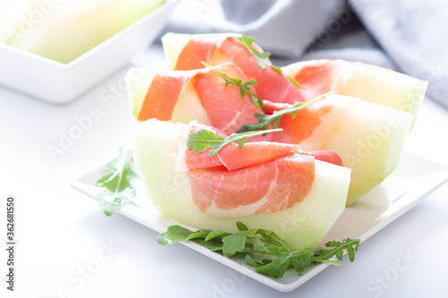 Traditional Italian or Spanish appetizer fresh melon with prosciutto or jamon and green herbs on white table, top view, light mediterranean cuisine dish